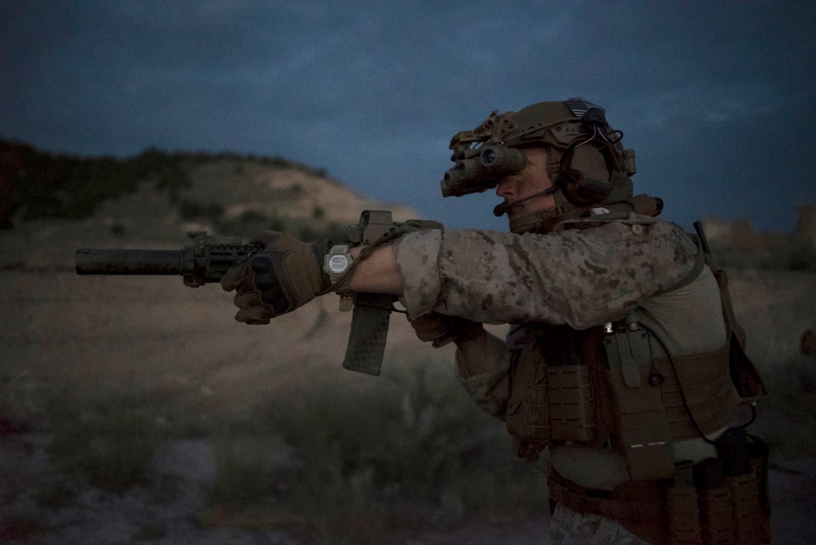 Soldier in camouflage aiming a rifle at dusk with night-vision goggles and tactical gear in a desert landscape.