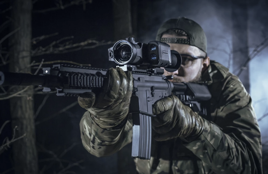 A soldier in camouflage gear aiming a rifle with a scope in a dark, wooded setting at night.