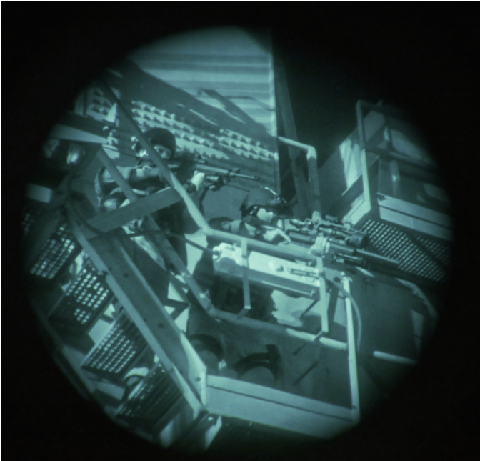 Night-vision view of a person with a rifle crouched on a metal catwalk, aiming through the sights in a dim industrial setting.