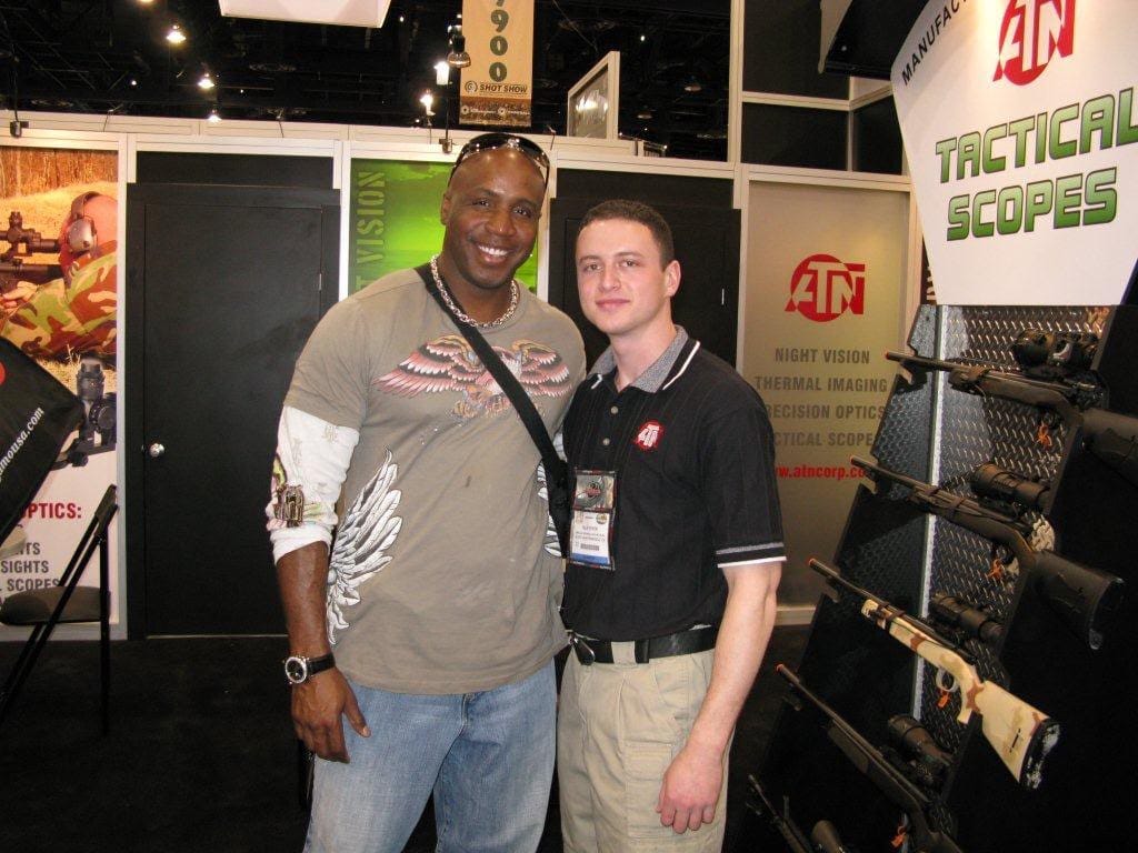 Two men pose at a trade-show booth with rifles on display behind them.