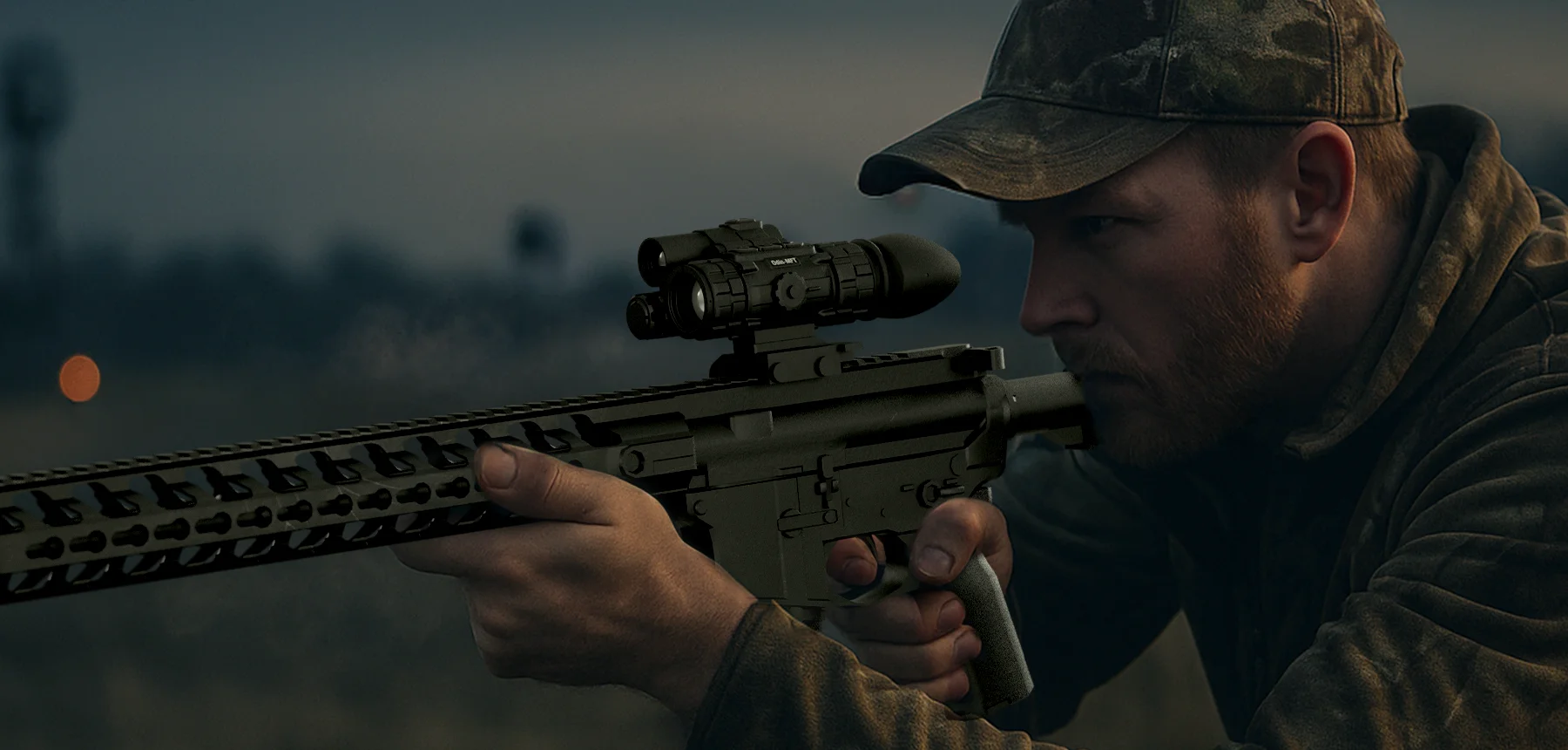 Man in a cap crouched and aiming a rifle with a scope outdoors at dusk.